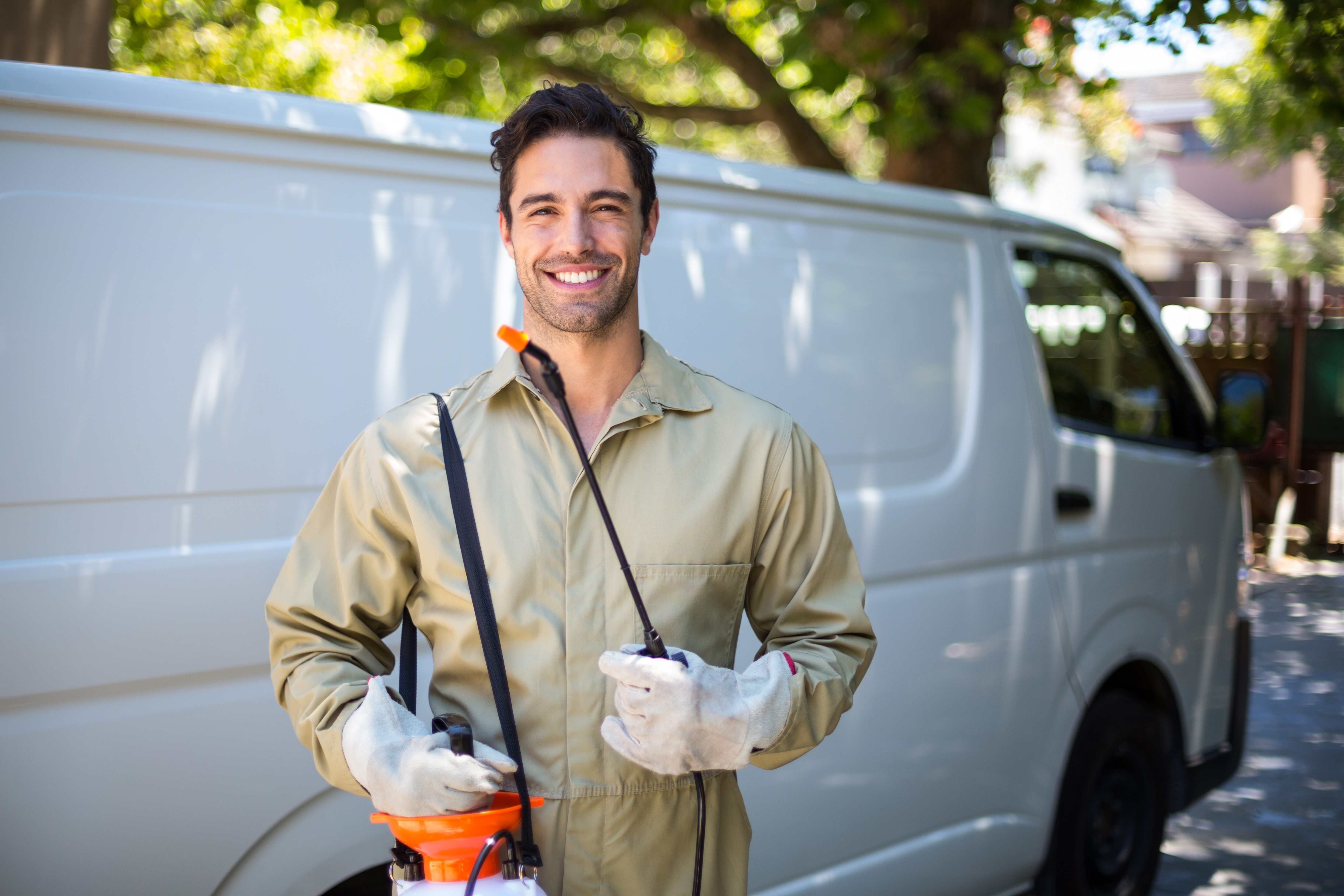 Pest control technician in blue uniform with protective gloves and orange sprayer backpack, loading equipment from a service van outside a residential home.