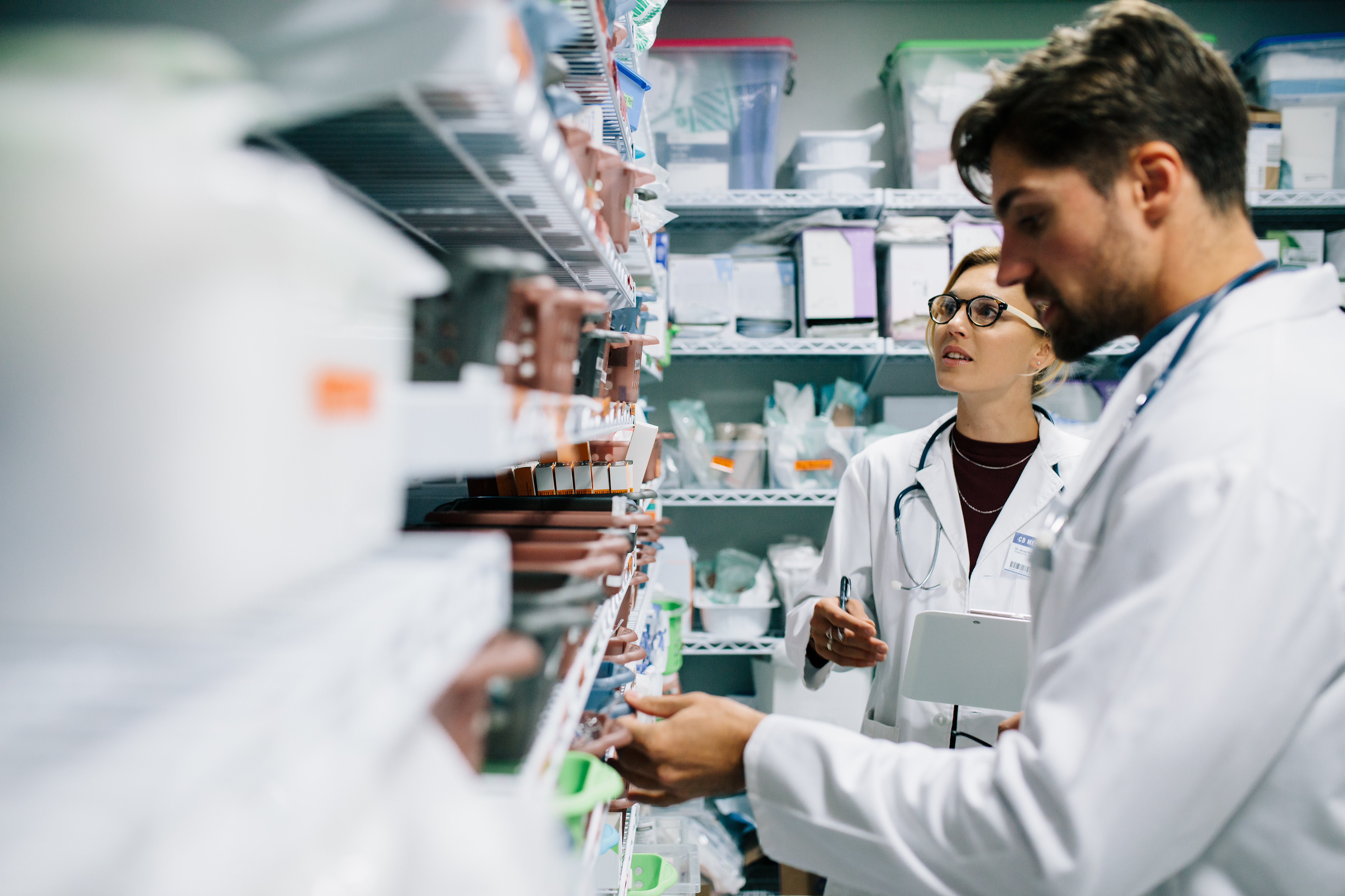 Two healthcare professionals in white lab coats review medical supplies in a storage room. The man examines items on a shelf while the woman holds a clipboard and takes notes, surrounded by neatly organized bins and containers.